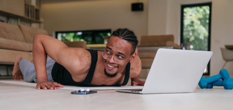 Training At Home. Sporty african american man doing the training while watching an online tutorial on a laptop, exercising in the living room, free space.