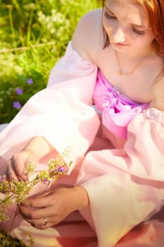 A young woman gently collects wildflowers while seated in a brightly lit meadow, adorned in a flowing pink dress during a warm afternoon.