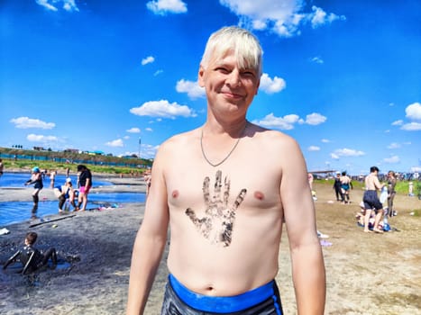 A man stands at the beach, proudly displaying a handprint design on his chest while friends swim nearby. The sunny day features clear blue skies and fluffy clouds.