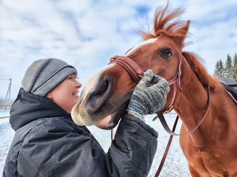 A woman is smiling while grooming a chestnut horse in a snowy area under a cloudy sky during winter.