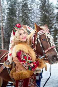 A beautiful Russian woman in vibrant dress poses with her horse, embodying Maslenitsa in a winter wonderland.
