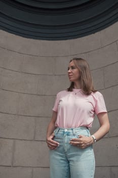 Young woman in a pink t-shirt and ripped jeans leans outdoor, looking thoughtful.