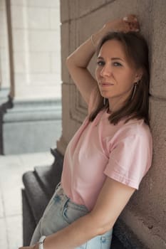 Young woman in a pink t-shirt and ripped jeans leans outdoor, looking thoughtful.
