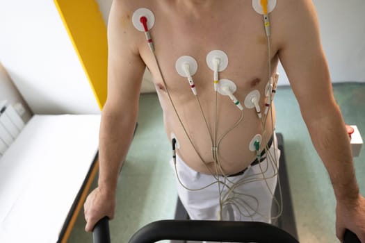 A middle-aged man is undergoing a medical stress test on a treadmill, connected to ECG electrodes for heart monitoring in a clinical environment.
