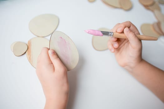 Child painting Easter wooden egg shapes with pink paintbrush on white surface.