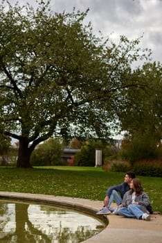 Autumn Serenity: Couple Relaxing by Reflective Pond Amidst Vibrant Fall Foliage and Ivy-Covered Architecture. Tranquil Garden Scene Captures Intimate Moment Against Backdrop of Colorful Autumn Leaves and Mirrored Water Surface. This enchanting autumn scene portrays a couple sharing a peaceful moment by a serene pond, surrounded by the rich hues of fall foliage. The pair sits comfortably on the grass, leaning against each other in quiet companionship as they gaze upon the reflective water. Vibrant red and orange leaves adorn the surrounding trees, creating a warm and inviting atmosphere. An ivy-covered building in the background adds a touch of timeless charm to the landscape. The pond's mirror-like surface perfectly captures the reflection of the couple and the colorful foliage, doubling the visual impact of autumn's beauty. This image embodies the essence of autumn tranquility, making it ideal for themes of romance, relaxation, and the changing seasons.