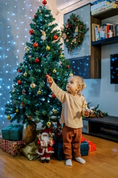 Little girl stands near the Christmas tree and points her finger up to the side. High quality photo