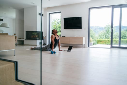 Training At Home. Sporty African American man doing the training while watching an online tutorial on a laptop, exercising in the living room, free space.