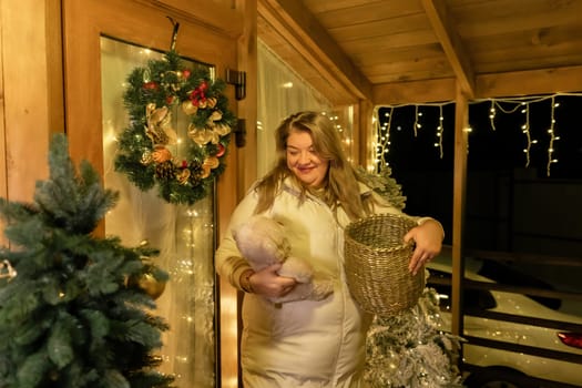 A woman in a white robe holds a wicker basket and smiles on a porch decorated with Christmas lights and a wreath.