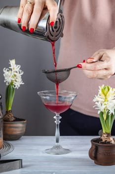 Bartender filtering red cocktail from shaker through strainer into a coupe glass, white hyacinth flowers on a gray background