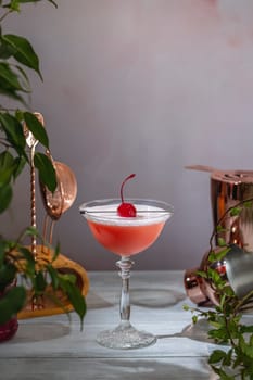 Refreshing pink cocktail with egg white foam, garnished with a maraschino cherry, served in a coupe glass on a wooden table with bar tools and plants