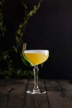 Bright yellow cocktail in a stemmed glass, garnished with a fresh green leaf and clothespin, resting on a dark wooden table