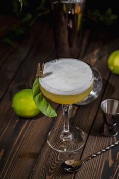 Vibrant yellow cocktail topped with white foam sits in a coupe glass on a dark wooden table, garnished with lime and leaf