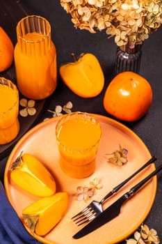 Glass of refreshing persimmon juice served with ripe persimmon fruits on a plate, decorated with dried flowers, creating a vibrant autumnal table setting
