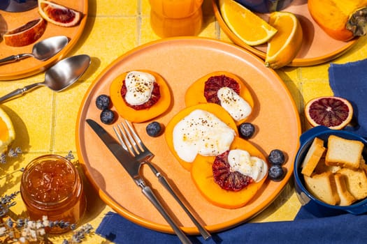 Delicious persimmon dessert with ricotta cheese, blood orange slices and blueberries, served on a vibrant table with orange juice, jam, and melba toast