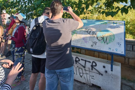 Crete.Greece - april 02, 2025: A group of tourists looking at a map of Heraklion outdoors, engaging with city details and planning their visit, with visible trees and a stone wall in the background.