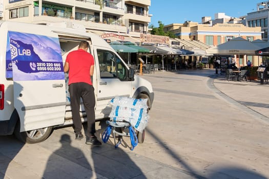 Crete.Greece - april 02, 2025: Ice delivery worker unloading bags from a white van in a busy plaza with restaurants and cafes, highlighting the daily operations of urban supply chains.