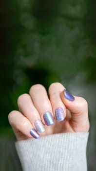 Skittles nails on hand. Colorful shiny manicure with different textures in light purple tones and lilac hue. Female hand with multicolored nails on green foliage background. Copy space. Vertical