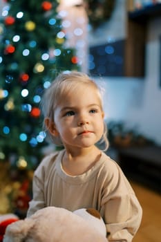 Little girl sits near a Christmas tree sparkling with colorful lights. Blur. High quality photo