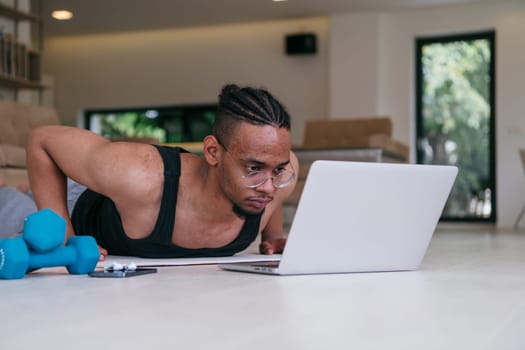 Training At Home. Sporty african american man doing the training while watching an online tutorial on a laptop, exercising in the living room, free space.