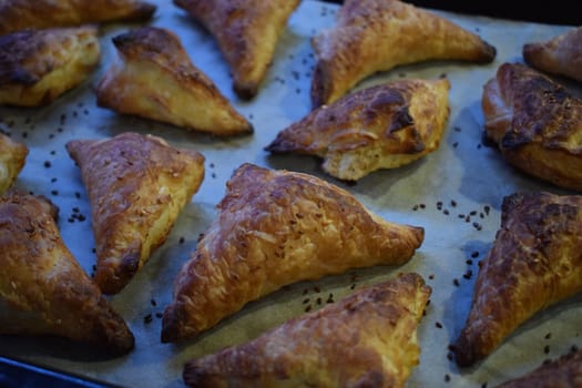 A baking tray filled with freshly baked golden brown triangular cakes. The cakes have a flaky, crunchy texture, are sprinkled with sezam seeds on top. They spread evenly on a sheet of parchment paper