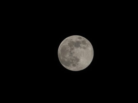 A full moon in the center, in a completely dark sky. Its illuminated surface is highlighted and visible in detail, with several craters and dark spots on a completely black background.