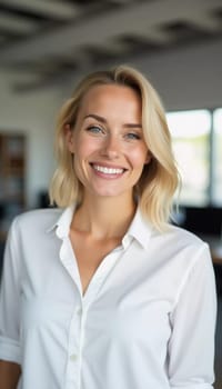 A cheerful blonde woman in a white blouse smiles warmly in a modern bank office. The well-lit space enhances her friendly demeanor, inviting clients for assistance.