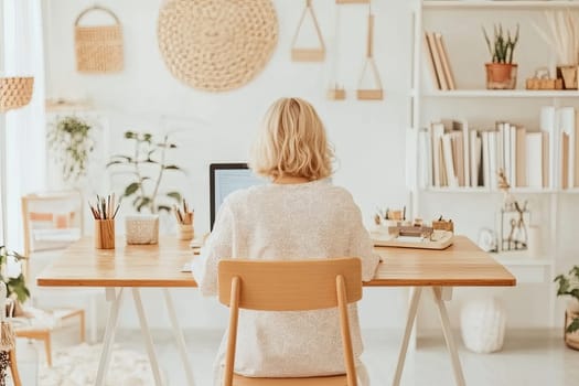 An elderly blonde woman sits at a wooden desk, focused on typing on her laptop.