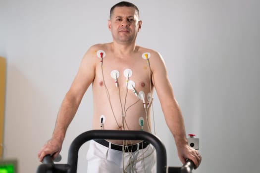 A middle-aged man is undergoing a medical stress test on a treadmill, connected to ECG electrodes for heart monitoring in a clinical environment.