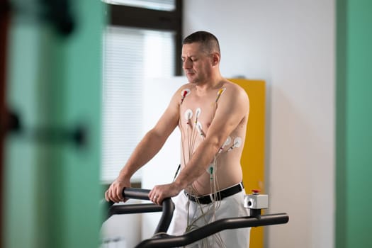 A middle-aged man is undergoing a medical stress test on a treadmill, connected to ECG electrodes for heart monitoring in a clinical environment.