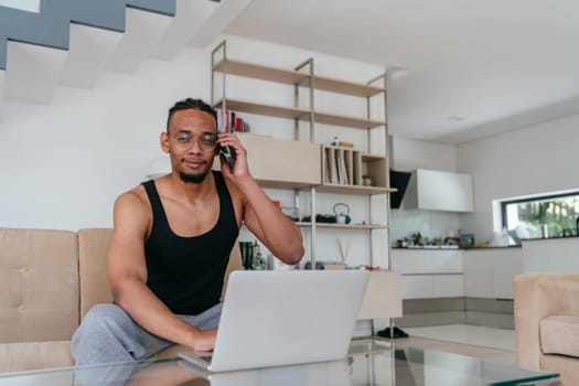 African American man in glasses sitting at a table in a modern living room, using a laptop for business video chat, conversation with friends and entertainment.