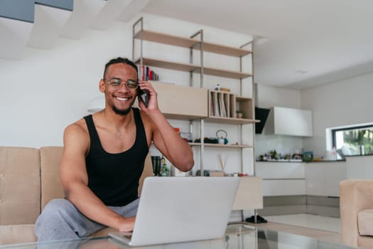 African American man in glasses sitting at a table in a modern living room, using a laptop for business video chat, conversation with friends and entertainment.