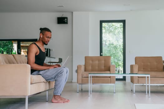 African American man in glasses sitting at a table in a modern living room, using a laptop for business video chat, conversation with friends and entertainment