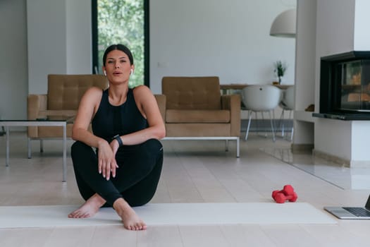 A young woman in athletic wear sits on a yoga mat in a spacious, modern living room after a workout. She looks relaxed, surrounded by exercise equipment including small dumbbells and a laptop.
