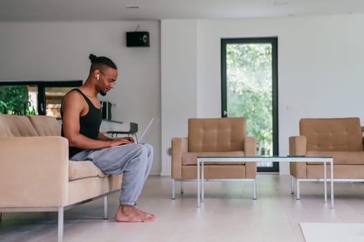 African American man in glasses sitting at a table in a modern living room, using a laptop for business video chat, conversation with friends and entertainment