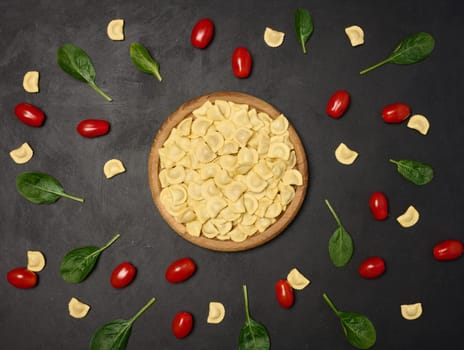 Uncooked tortellini pasta with fresh cherry tomatoes and spinach leaves on a dark, textured surface. Flat lay 