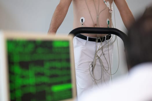 A middle-aged man is undergoing a medical stress test on a treadmill, connected to ECG electrodes for heart monitoring in a clinical environment.