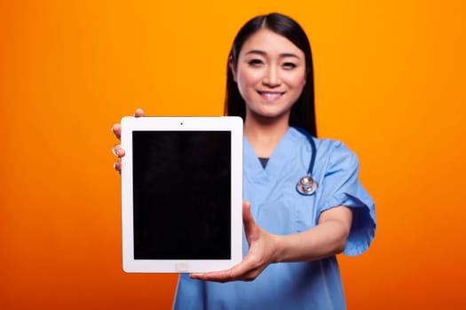 Asian doctor holding a digital device displaying black screen, mockup template. Female nurse in blue scrubs, presenting blank tablet, perfect for medical campaigns, set against an orange background.