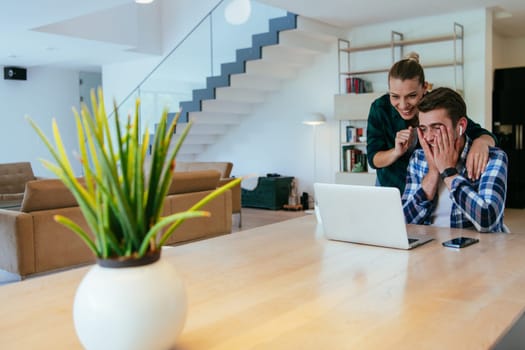A young married couple is talking to parents, family and friends on a video call via a laptop while sitting in the living room of their modern house