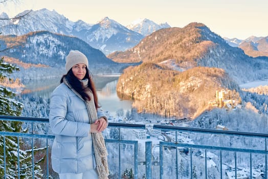 Captivating lifestyle portrait of a fashionable young woman in warm winter attire checking her mobile phone while exploring a scenic mountain viewpoint. The scene combines modern technology with pristine nature, featuring the subject in a light gray puffer coat and beige accessories against a breathtaking backdrop of snow laden evergreens, distant peaks, and a charming village nestled below, all illuminated by soft golden sunlight, creating perfect winter travel and outdoor lifestyle content. Young Woman Using Smartphone in Winter Mountain Landscape While Standing on Viewpoint Above Snow Covered Village in Golden Evening Light