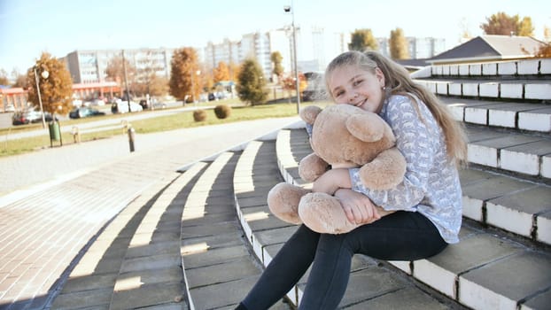 Young blonde and smiling teen girl with teddy bear toy outdoors.