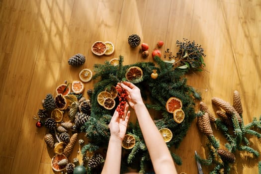 Female hands hold a sprig of red berries over a Christmas wreath lying on the floor. Top view. High quality photo