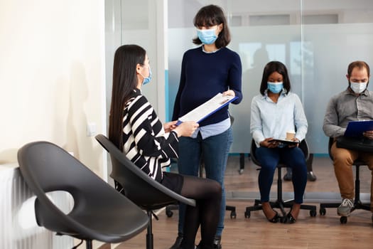 Asian woman seated in lobby area, giving cv resume to pregnant recruiter, ready for employment meeting. Female job applicant and caucasian expectant lady hold clipboard, discussing interview process.