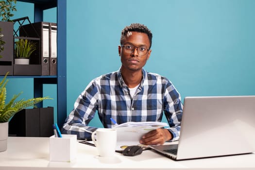 African american man with spectacles sitting at the table with documents and laptop, doing research work. Portrait of a black male entrepreneur holding papers and looking at the camera.