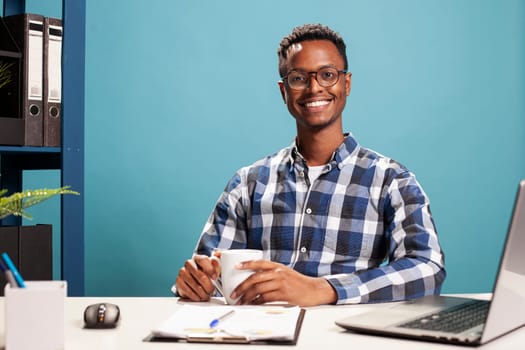 Young black man with coffee cup in his hands, is sitting at his desk and smiling at the camera. African american male freelancer seated at table with clipboard and a laptop, ready for work.