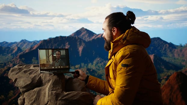 Workaholic explorer meeting with colleagues on online video call, enjoying a strong signal high speed internet on top of the mountain. Young hiker CEO with dependency. Camera A.