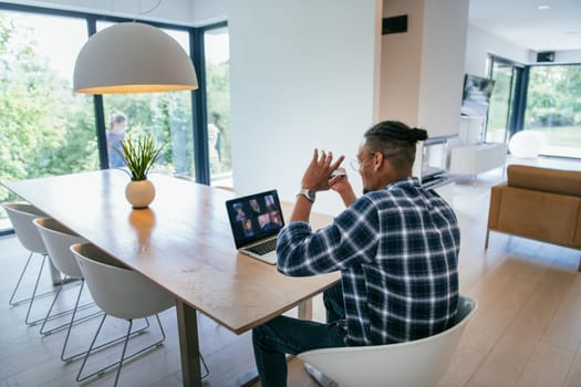 African American businessman in glasses sitting at a table in a modern living room, using a laptop and smartphone for business video chat, conversation with friends and entertainment