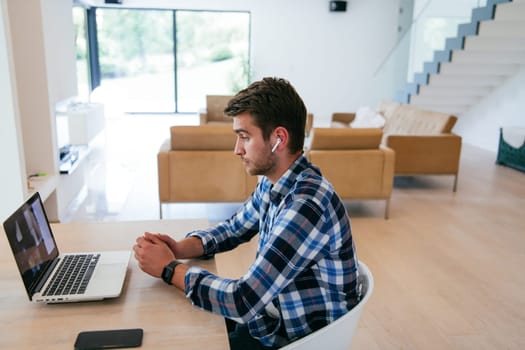 A freelancer sitting at a table in a modern living room, with headphones using a laptop for business video chat, conversation with friends and entertainment.