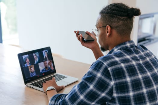 African American businessman in glasses sitting at a table in a modern living room, using a laptop and smartphone for business video chat, conversation with friends and entertainment