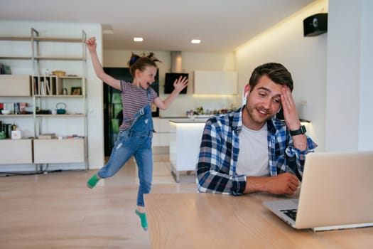 Work from home. Daughter interrupting her father while he is having a business online conversation on his laptop while sitting in the modern living room